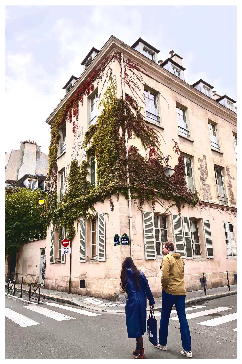 a building from le marais, paris is clearly visible. there is a couple walking on the street. the woman has a blue coloured trench coat and the man is wearing a beige jacket.